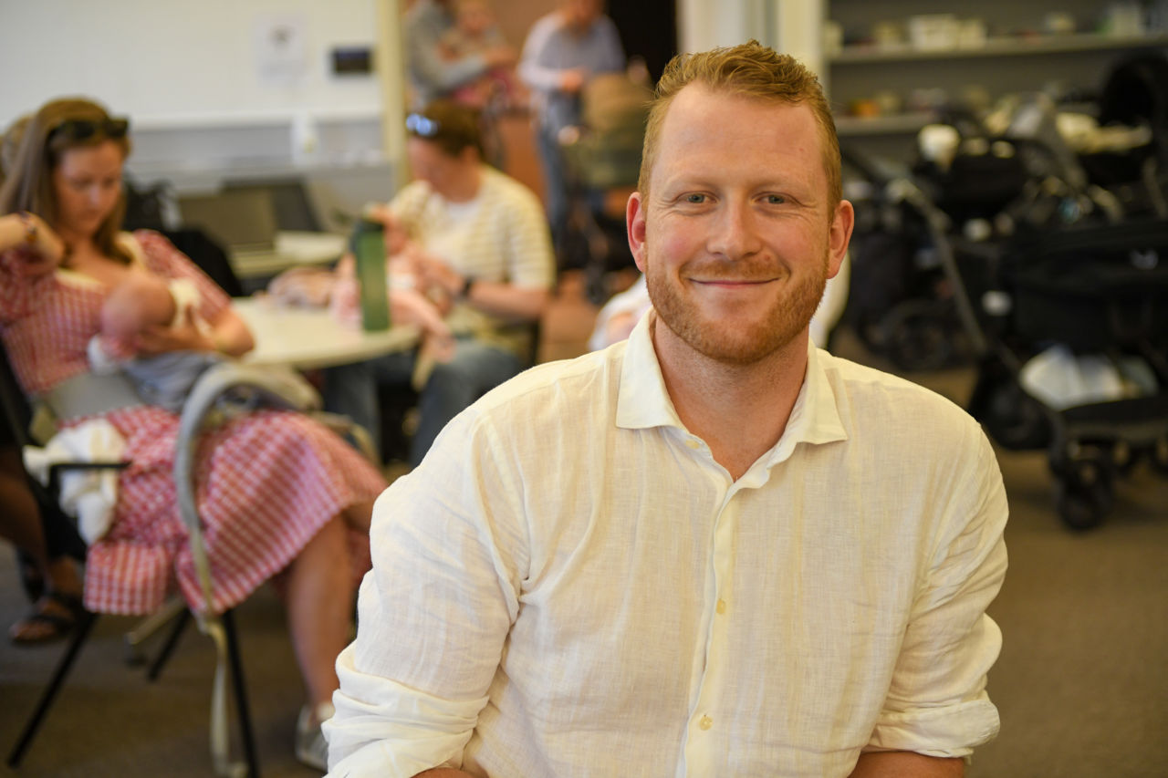 Portrettbilde av en smilende mann med flere mødre og ferdre med babyer i bakgrunnen.