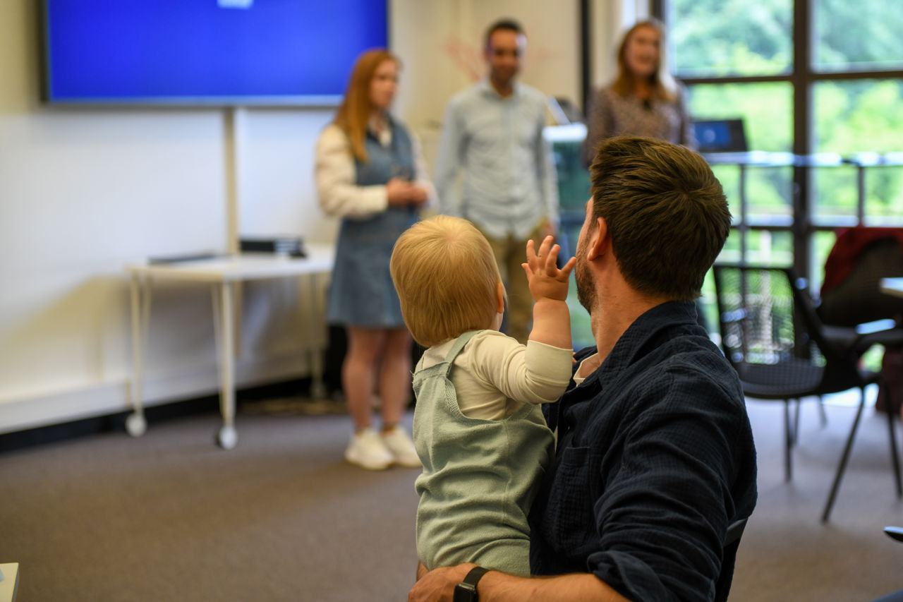Far og barn fotografert bakfra mens de ser på tre personer som snakker foran dem.