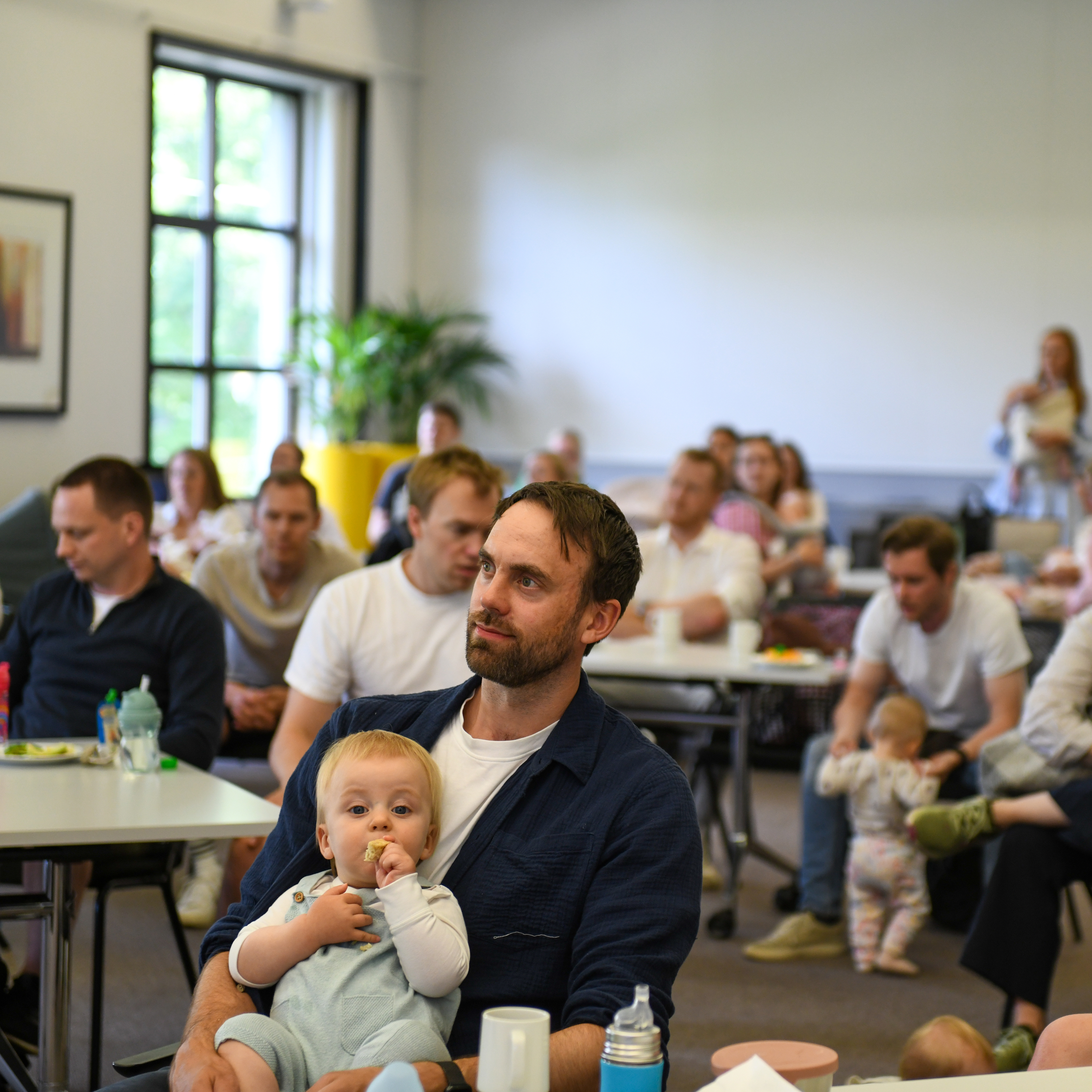A male NHH alumni on parental leave with his baby in his arms listening to a presentation,.