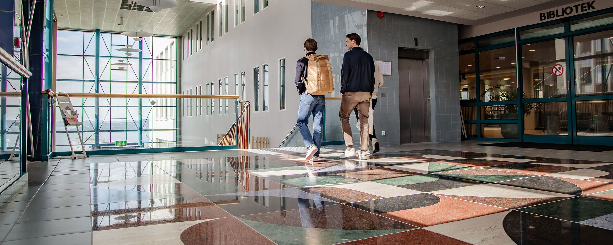 Two students wearing backpacks are walking passed  the library towards a staircase at NHH's campus.