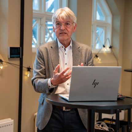 An older man with grey hair and glasses, wearing a grey blazer over a white shirt, gestures with both hands while standing behind a laptop at a high table during a presentation.
