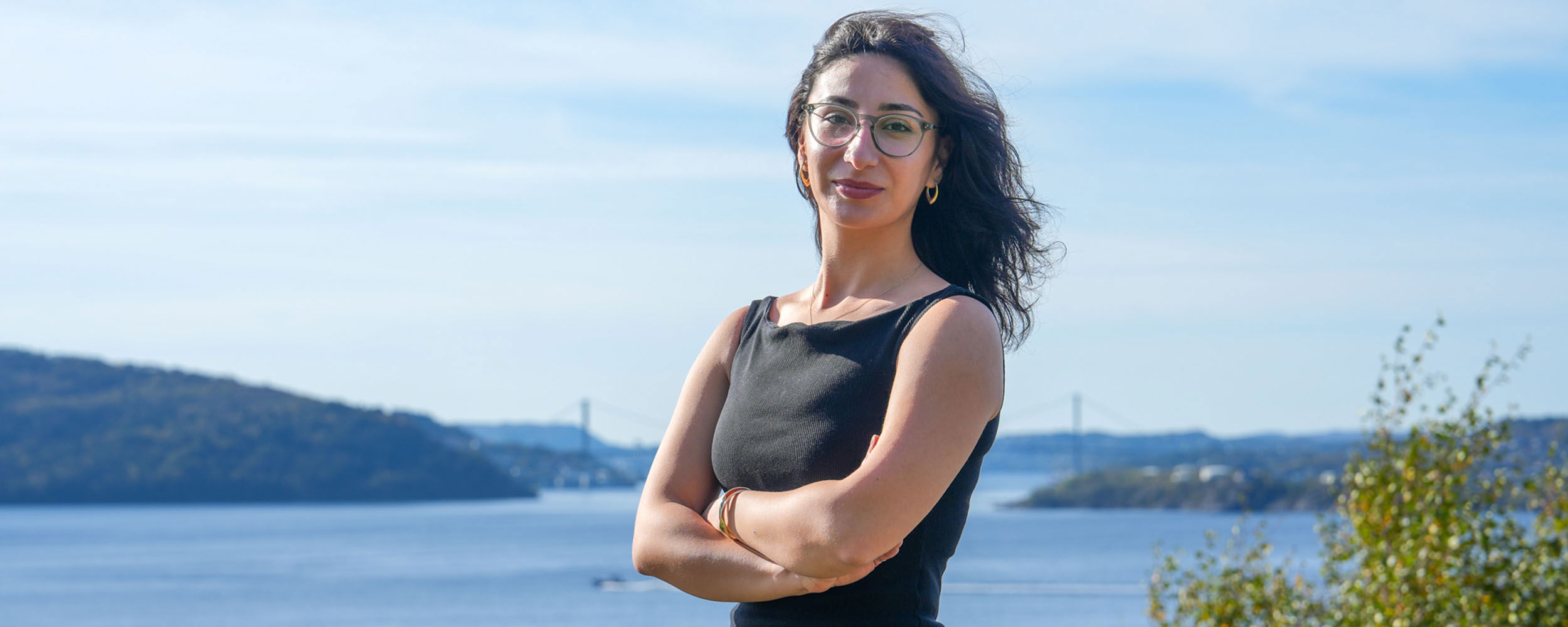 Portrait of a smiling woman wearing glasses, standing outdoors with the sea in the background on NHH’s campus.