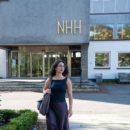 A woman wearing glasses, a black top, a dark skirt and a shoulder bag walks across the campus of the Norwegian School of Economics (NHH), with the NHH sign visible on the building behind her.