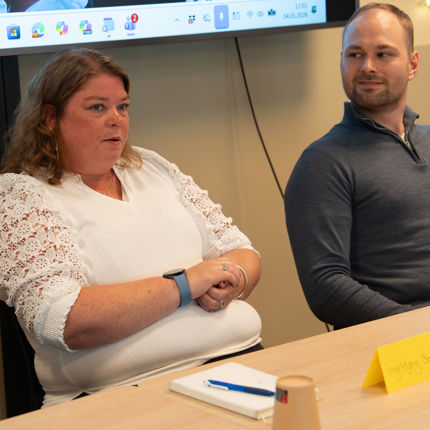 A woman in a white lace-sleeved top speaks during a meeting while a man in a blue pullover listens, seated at a table with a notebook and a yellow name tent.
